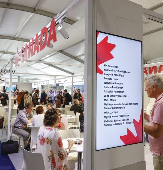A large, white trade-show booth with a dozen tables, each holding a meeting. At the front of the pavilion is a screen decorated with maple leaves, listing companies that are partaking in the meetings. A red "Canada" sign adorns the top of the booth.