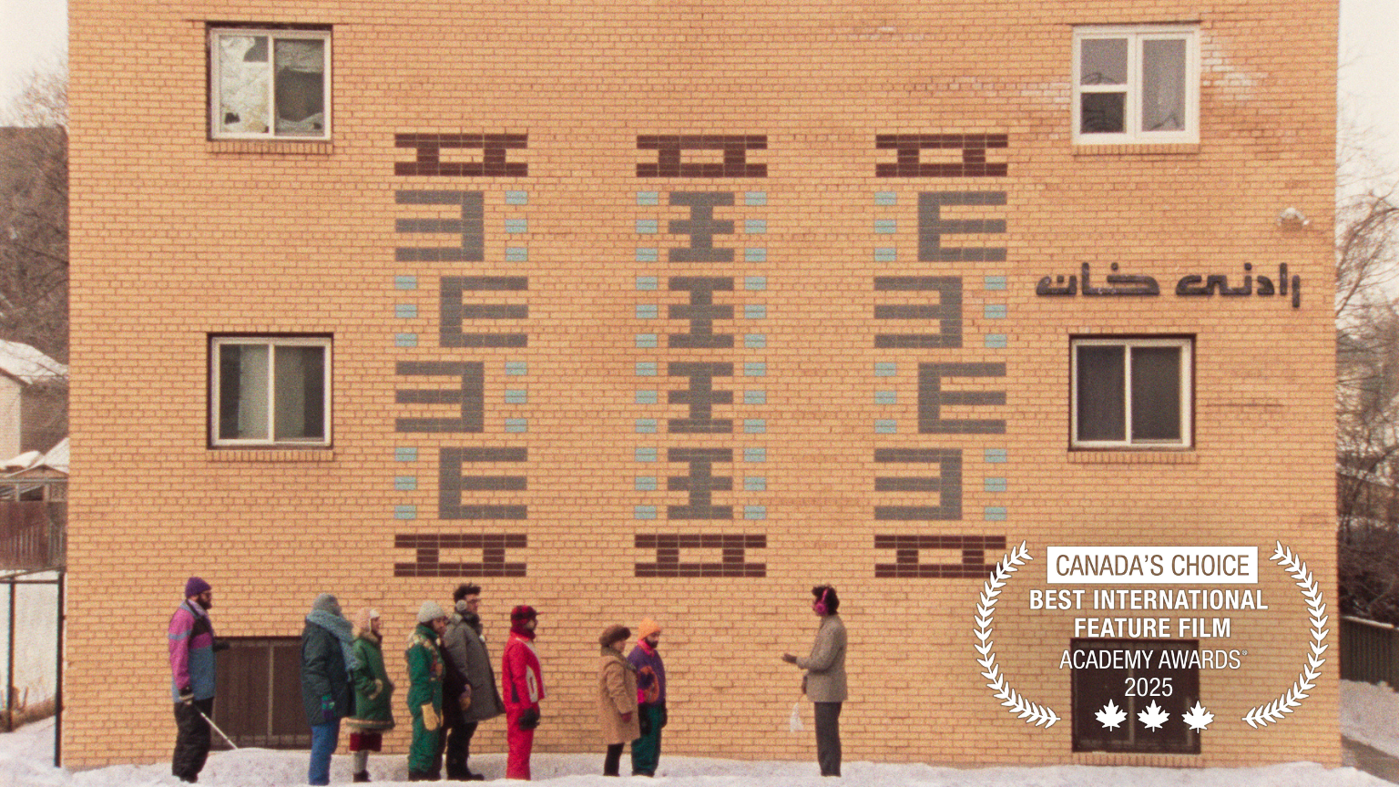 A still from the film Universal Language directed by Matthew Rankin. It is winter. A tour group dressed in bright colours stand in front of a beige brick building, while a tour guide with red earmuffs explains the significance of the spot. In the bottom right-hand corner, laurels that say "Canada's Choice. Best International Feature Film. Academy Awards 2025".